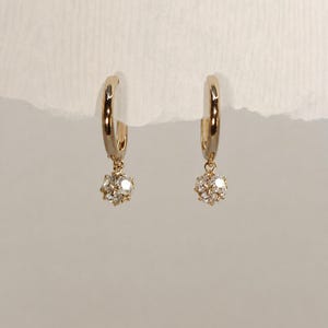 May include: A pair of gold hoop earrings, each featuring a small, dangling spherical charm encrusted with multiple clear, faceted stones. The hoops have a polished, reflective finish and the charms are attached by a small, circular jump ring.