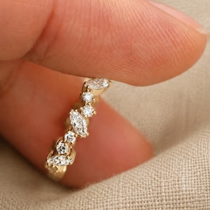May include: A gold ring featuring a curved band set with a series of alternating marquise-cut and round-cut clear stones. The stones are arranged in a cluster pattern along the top portion of the band, creating a delicate and shimmering appearance.
