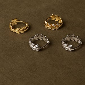 May include: Four hoop earrings arranged in a group. Two earrings are gold-toned metal with a leaf-shaped design, and two are silver-toned metal with a leaf-shaped design. Each earring features small, clear, sparkling stones set along the leaves.