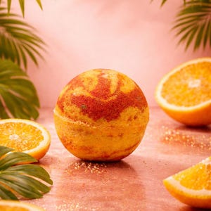 Puede incluir: Una bomba de baño naranja con un patrón rojo, rodeada de rodajas de naranja y hojas verdes. El fondo es rosa con purpurina, creando un ambiente de spa. Perfecto para un baño relajante.
