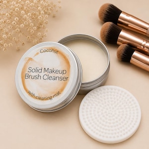 May include: A round silver tin containing solid makeup brush cleanser with a label reading Coconut Solid Makeup Brush Cleanser with cleaning mat. Beside the tin is a white circular silicone cleaning mat and three makeup brushes with rose gold handles.
