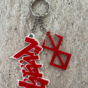 Op de afbeelding: Een sleutelhanger met twee rode acryl bedels bevestigd aan een zilverkleurige metalen ring. Eén bedel toont rode Japanse katakana-tekens met een witte omlijning, terwijl de andere bedel een rood geometrisch symbool is dat lijkt op een merk.