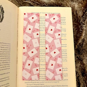 Peut inclure: Deux marque-pages rectangulaires avec un fond rose clair orné d'un motif répétitif d'enveloppes blanches scellées par des cœurs rouges et de petites étoiles roses à quatre branches, posés verticalement sur les pages d'un livre.