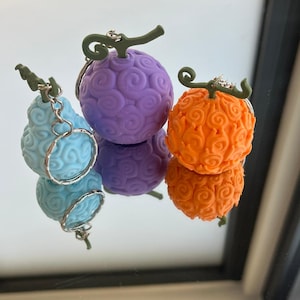 Peut inclure: Trois porte-clés en forme de fruits, de couleurs bleu clair, violet et orange. Chaque porte-clé a une tige verte et un anneau argenté. Ils sont présentés sur une surface réfléchissante.