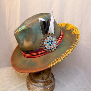 May include: A distressed olive green fedora hat featuring a wide brim with yellow stitched edges. The crown is adorned with a circular beaded medallion in blue, orange, and black, two feathers, and a layered band of red and orange cord trim.