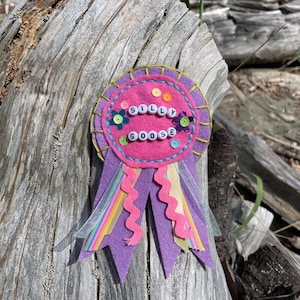 May include: A handmade pink and purple felt badge with the words "SILLY GOOSE" spelled out in white letter beads. The badge is decorated with colourful sequins, flower-shaped beads, and ribbons.
