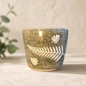 以下が含まれることがあります： 白の植物の葉の模様が施された、グレーとグリーンの斑点模様の釉薬がかかった陶器のカップ。外側の表面には、大きなシダの葉のデザインと、それより小さなモンステラの葉のモチーフが描かれており、滑らかで丸みを帯びた縁で仕上げられています。