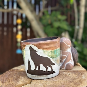 Könnte beinhalten: Handgefertigte Keramiktasse mit einer Silhouette eines heulenden Wolfs auf weißem Grund. Die Tasse hat braune, grüne und weiße Farbbänder. Eine kleine Schildkrötenfigur steht daneben.