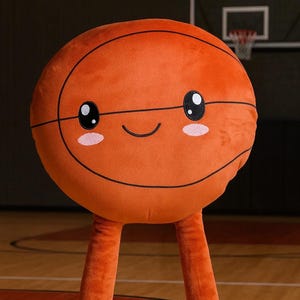May include: An orange plush basketball with a smiling face, black lines, and blush details. It has long orange legs and is wearing light blue sneakers with white laces. The background shows a basketball court.