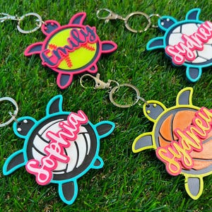 May include: Four turtle-shaped keychains with sports-themed designs. One features a softball, another a volleyball, and two with basketballs. Each keychain has a name in a cursive font, and a metal clasp.