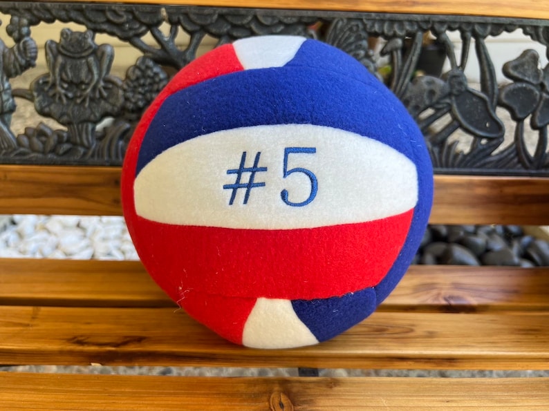 May include: A soft volleyball in red, white, and blue. The ball features a white panel with the embroidered number "#5" in blue. The ball is resting on a wooden bench with decorative carvings.