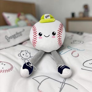 May include: A plush baseball character with a smiling face, red stitching, and a small green hat. It has gray legs and wears white and navy blue shoes. The baseball is on a white bedspread with baseball-themed illustrations.