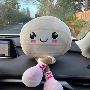May include: A plush volleyball toy with a smiling face, light brown stripes, and pink and white legs. The toy is sitting on a car dashboard, with a road and trees visible in the background.