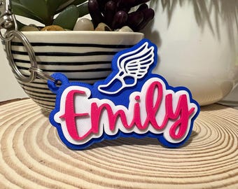 Può includere: Un portachiavi blu, bianco e rosa con il nome "Emily" in corsivo. Un disegno di ali bianche è sopra il nome. Il portachiavi è attaccato a un anello di metallo argentato.