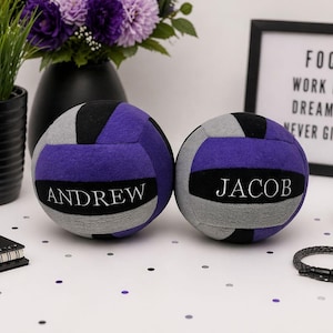 May include: Two personalised volleyballs in purple, grey, and black, each with a name in white. The volleyballs are on a white surface with confetti, near a framed sign that reads "FOCUS WORK HARD DREAM BIG NEVER GIVE UP."