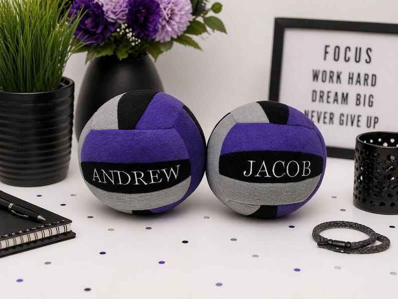 May include: Two personalised volleyballs in purple, grey, and black, each with a name in white. The volleyballs are on a white surface with confetti, near a framed sign that reads "FOCUS WORK HARD DREAM BIG NEVER GIVE UP."