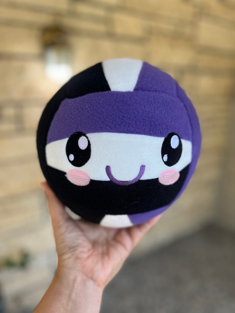 May include: A plush volleyball toy with a smiling face. The ball is in black, white, and purple. The face has large black eyes, pink cheeks, and a purple stitched smile. The toy is held in a hand.