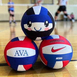 May include: Three volleyballs stacked. The top ball is blue, white, and black with a cartoon face. The bottom two balls are red, white, and blue. One has the name "AVA" and the other has a Nike logo and the text "HYPER SPIKE".