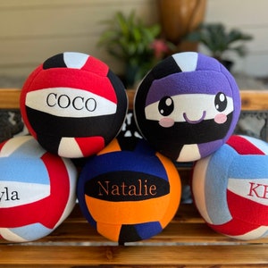 May include: Several colourful volleyballs, each with a name printed on them. The balls are in shades of red, blue, and purple. Names include "COCO", "Khyla", "Natalie", and "Kenzie". One ball has a cartoon face. They are arranged on a wooden bench.