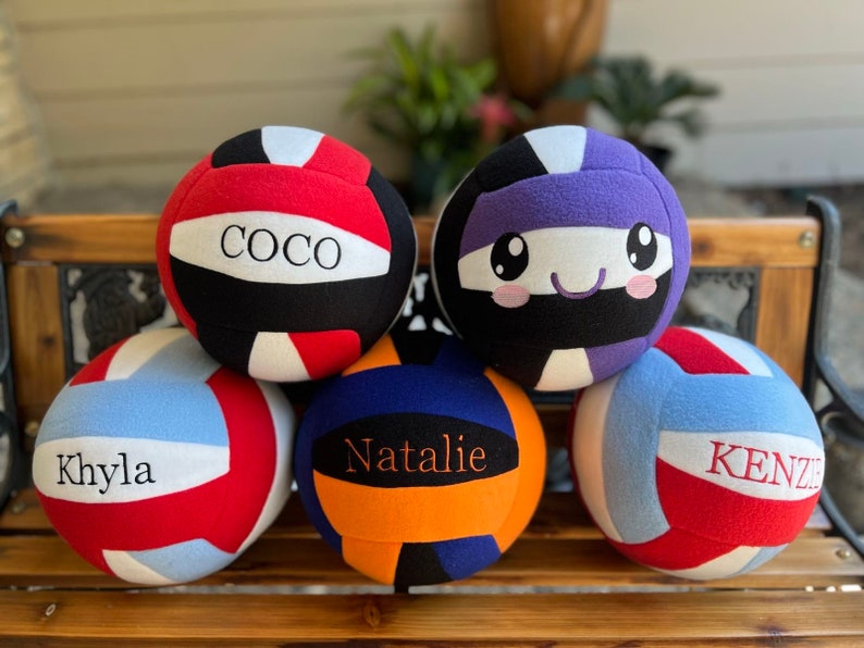 May include: Several colourful volleyballs, each with a name printed on them. The balls are in shades of red, blue, and purple. Names include "COCO", "Khyla", "Natalie", and "Kenzie". One ball has a cartoon face. They are arranged on a wooden bench.