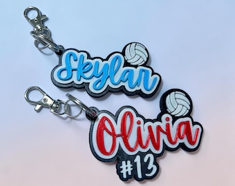 May include: Two personalized volleyball keychains. One keychain has the name "Skylar" in blue, and the other has the name "Olivia" in red with the number "13". Each keychain includes a volleyball graphic.