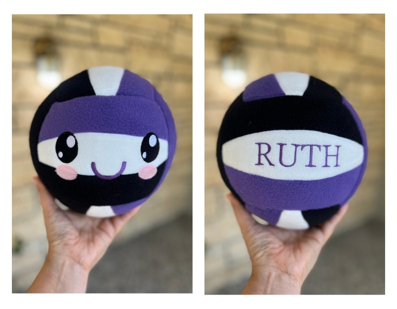 May include: A soft volleyball in black, white, and purple, with the name "RUTH" embroidered on one side. The other side features a cartoon face with large eyes and a smiling mouth. The ball is held in a person's hand.