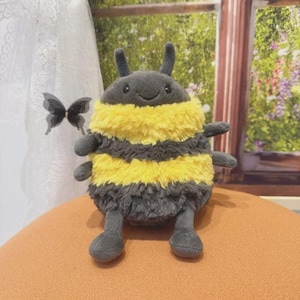 May include: A plush bumblebee toy with a soft, textured body. The bee features a gray head and legs, with a yellow and black striped body. It has small antennae and a smiling face. The toy is designed to resemble a bumblebee.