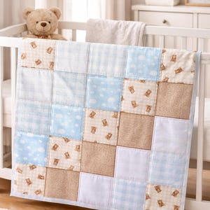 Könnte beinhalten: Eine Patchwork-Babydecke mit Quadraten aus blauem, weißem und beigem Stoff. Einige Quadrate zeigen Teddybären-Prints, Wolken oder Vichy-Muster. Die Decke ist über einem weißen Babybett drapiert.