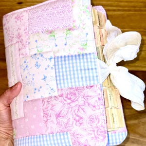 Op de afbeelding: Een gewatteerde tijdschriftomslag in roze, blauw en wit, met een patchworkontwerp met bloemen- en ruitpatronen. Het tijdschrift is vastgebonden met een wit lint en heeft gelabelde tabbladen.