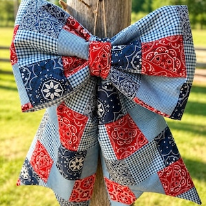 May include: A large decorative bow made from a patchwork of fabric squares featuring red, white, and navy blue bandana patterns mixed with light blue and white gingham checks. The bow is constructed with multiple fabric panels and a central knot.