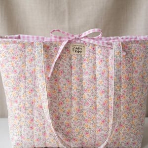 Könnte beinhalten: Eine gesteppte Tragetasche mit einem Blumenmuster in Rosa, Gelb und Weiß. Die Tasche hat ein rosa Vichy-Futter und eine passende Schleife. Sie hat lange Träger zum Tragen und ein kleines Etikett mit der Aufschrift "Handmade by Julia Ruby".