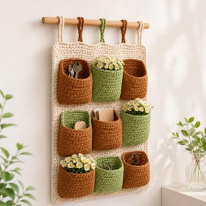 May include: A handmade, cream-colored crochet wall organizer with nine pockets in shades of brown and green. The pockets hold keys, small notecards, and small white flowers. The organizer hangs from a wooden dowel with cream, brown, and green hanging loops.