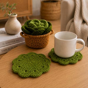 May include: A wooden table displays a white mug on a green crochet coaster, and a second coaster. A brown crochet basket holds a green crochet plant. A small vase with a plant and books are in the background.