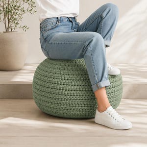 May include: A round, sage green crocheted pouf ottoman with a thick, textured yarn construction. The pouf is circular in shape and features a consistent, repeating stitch pattern throughout its exterior surface, providing a dense and firm appearance.