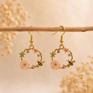 May include: A pair of gold-colored hoop earrings. Each earring features a floral design with pink and green flowers, small crystals, and a black and yellow bee. The earrings are hanging from a wooden branch.