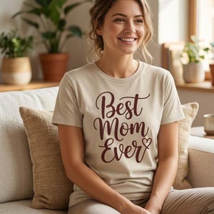 Op de afbeelding: Beige T-shirt met de tekst "Best Mom Ever" in een donker bordeauxrood script. Het shirt wordt gedragen door een persoon die op een bank zit. De persoon glimlacht. De achtergrond bevat planten in potten en een decoratief kussen.