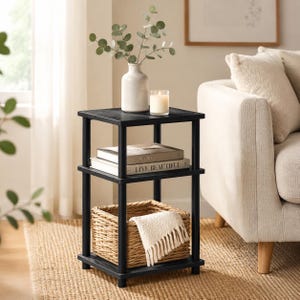 May include: A three-tier black side table with four cylindrical legs. The top shelf holds a white ceramic vase with eucalyptus branches and a glass candle. The middle shelf holds two books. The bottom shelf holds a woven basket with a white throw.
