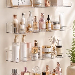 May include: Clear acrylic shelves displaying various beauty products and decorative items. The top shelf holds a framed print with the text "BE HAPPY EVERY DAY", a candle, and a small figurine. Other shelves showcase perfumes, lotions, and skincare products.
