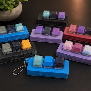 May include: Multiple rectangular plastic stands, each holding three mechanical keyboard keycaps in various colors and finishes, including glitter, matte, and translucent options. Each stand features the embossed text CUSTOMIZE on the front side.
