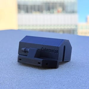 Op de afbeelding: Een zwart 3D-geprint apparaat met de tekst "Cyberswipe" aan de zijkant. Het apparaat heeft een rechthoekige vorm met een gebogen bovenkant en een uitstekende arm. Het ligt op een getextureerd oppervlak.