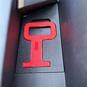 May include: A matte black interior car component featuring a recessed rectangular area with a bright red, T-shaped insert. The red insert includes a small, downward-pointing arrow graphic embossed in the center of its vertical section.