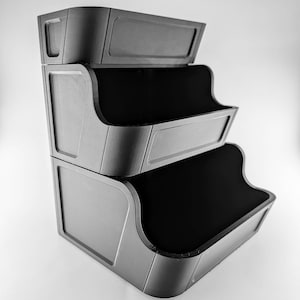 May include: A three-tiered, grey storage unit with a contemporary design. Each tier features a curved front and a rectangular panel. The interior of each tier is black. The unit is made of a solid material, suitable for organising items.