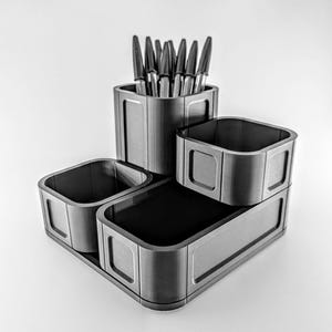 Pode incluir: Um organizador de secretária cinzento com vários compartimentos. O organizador inclui um porta-canetas alto cheio de canetas, juntamente com várias bandejas quadradas e retangulares. O design é moderno e minimalista, adequado para escritório ou casa.