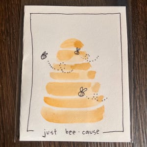 May include: A watercolour art print showing a stylised beehive in orange and yellow, with two bees and dotted flight paths. The phrase "just bee cause" is written below the beehive in black ink, within a black border.