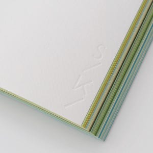 May include: A stack of white paper sheets with a textured surface, featuring the embossed letters S, K, and Y arranged vertically. The edges of the paper stack reveal thin, alternating layers of green, yellow, and blue colors.