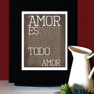 May include: A framed print with the words "El Amor" in large, distressed white letters, with a list of qualities of love in Spanish. A white pitcher and flowers are in the foreground.