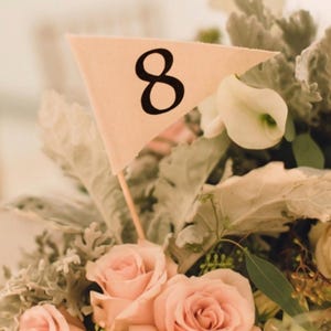 May include: A triangular cream-colored fabric flag featuring the black number 8 printed on its surface. The flag is attached to a thin wooden stick and is positioned among a floral arrangement of light pink roses and dusty miller foliage.