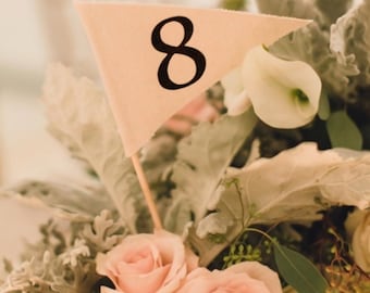 May include: A triangular cream-colored fabric flag featuring the black number 8 printed on its surface. The flag is attached to a thin wooden stick and is positioned among a floral arrangement of light pink roses and dusty miller foliage.