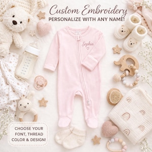May include: A light pink baby sleeper with the name "Sophia" embroidered on the chest, surrounded by baby items. The image includes a bottle, pacifier, socks, a wooden elephant teether, and a blanket. Text reads "Custom Embroidery" and "Choose your font, thread color & design!"