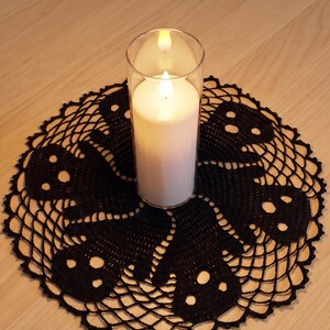 Puede incluir: Un tapete circular de ganchillo negro con un patrón repetido de fantasmas y bordes de malla calada. Un cilindro de vidrio transparente que contiene una vela de pilar blanca se sitúa en el centro. El texto "Halloween Ghost Doily Crochet Pattern" es visible.